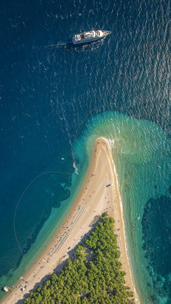 Aerial view of a narrow sandy beach stretching into deep blue water, with a large yacht rental anchored nearby and dense green trees covering part of the shore. The sea shows varying shades of blue and turquoise.