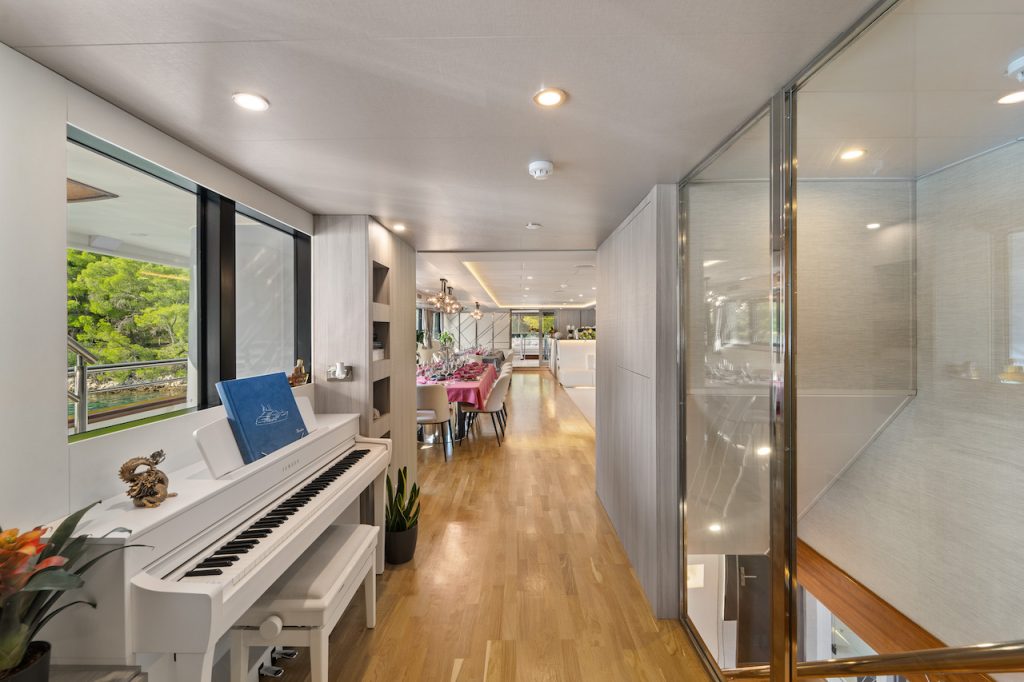 Modern hallway with a white piano on the left, wooden floor, and glass wall on the right evokes the elegance of a yacht charter. At the corridor’s end, a dining area set for a meal invites you in, while plants add a fresh touch of greenery.