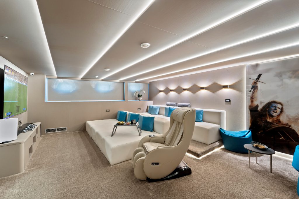 A modern home theater room with a large screen showing a sports game, tiered seating with white sofas and blue pillows, a reclining chair, ambient ceiling lights, and a wall mural of a yacht charter battle scene.