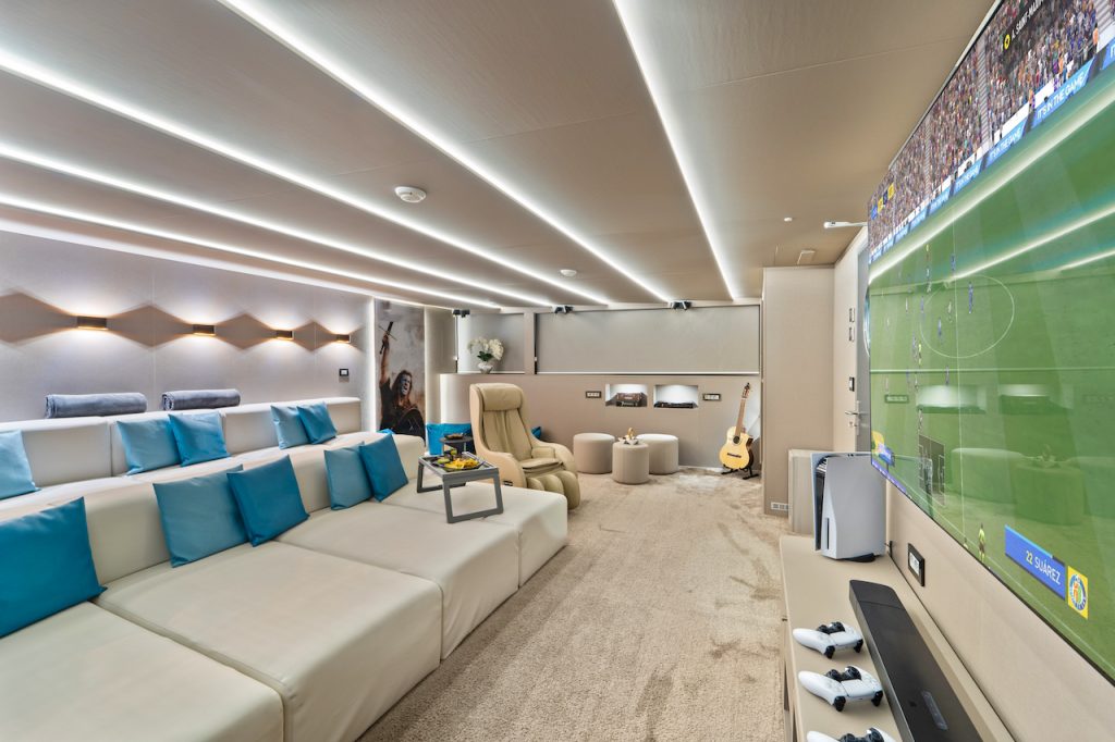Modern home theater with sectional seating, blue accent pillows, a large flat-screen TV showing a soccer game, gaming controllers, lounge chairs, guitar, soft lighting, and beige carpeting—perfect for planning your next yacht charter or relaxing after a day on the water.