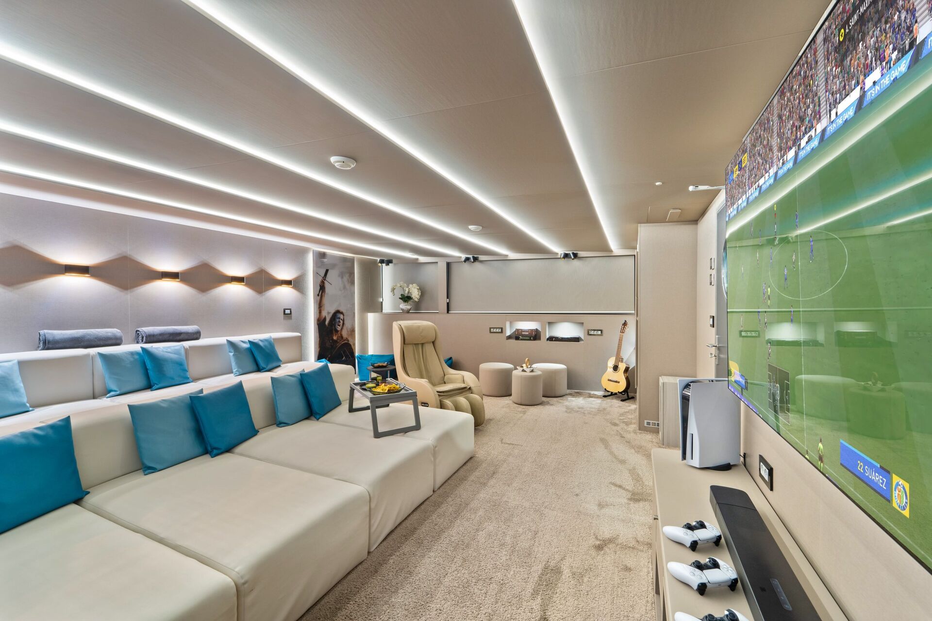 A modern home theater room with a large screen showing a soccer game, white sectional seating with blue pillows, lounge chairs, a guitar, soft lighting, and gaming controllers on a console—perfect for relaxing after a day on your yacht charter.