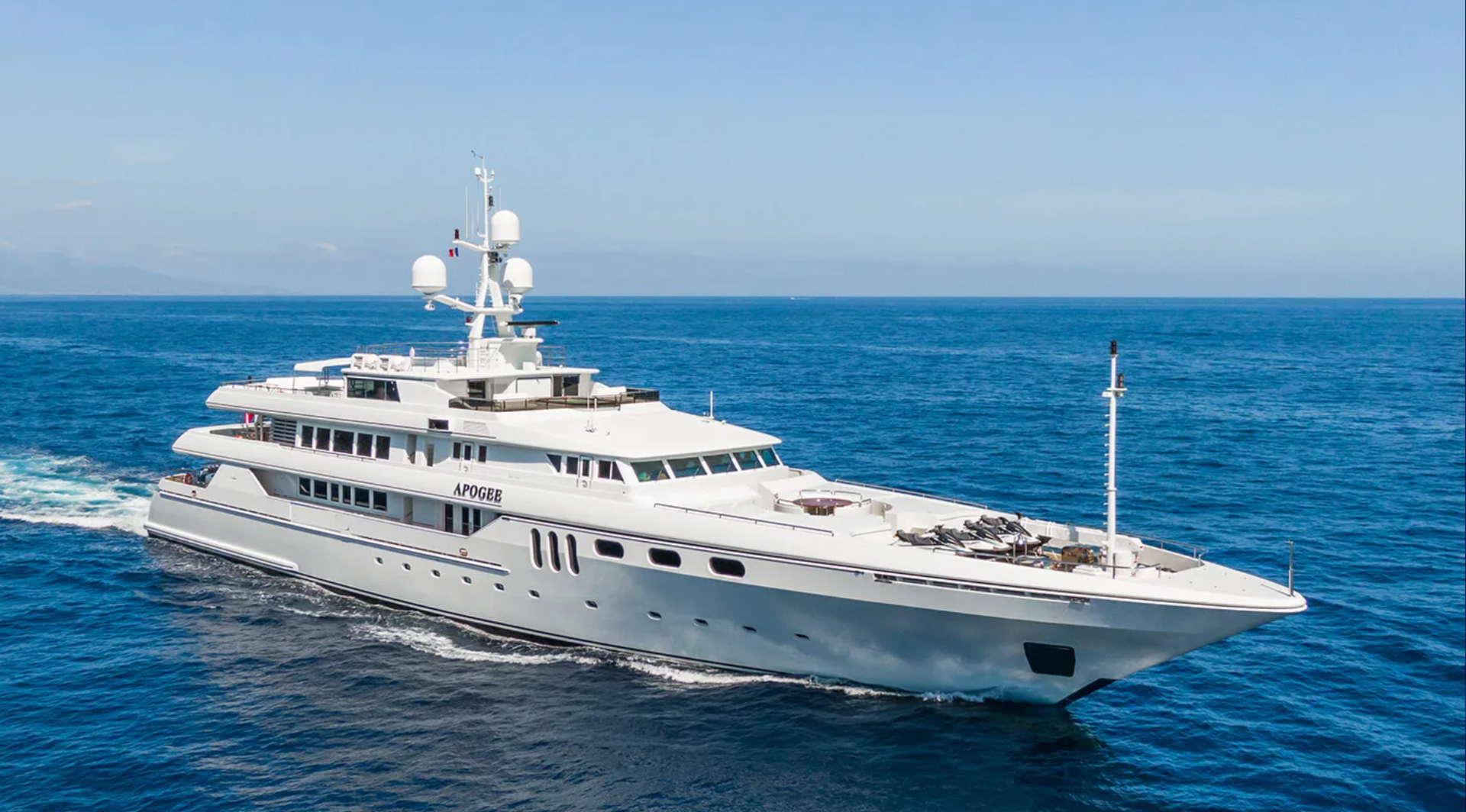 A large white luxury yacht sails across a calm blue ocean under a clear sky, leaving a trail of white waves behind it.
