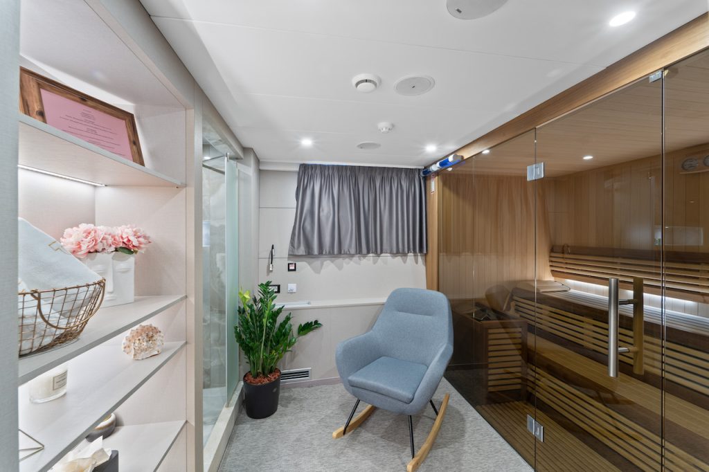 A modern spa room with a light blue rocking chair, a potted plant, built-in shelves, and a glass-walled wooden sauna—perfect for relaxing after an exclusive yacht charter. The white walls, gray curtains, and soft lighting create a tranquil retreat.
