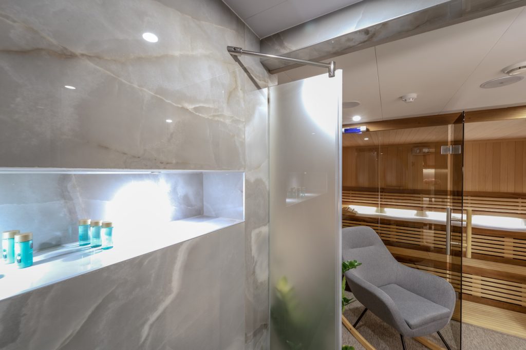 A modern spa room with marble walls, a frosted glass door, a built-in shelf holding toiletries, a gray lounge chair, and a wooden sauna—perfect for unwinding after a luxurious day on your yacht charter.