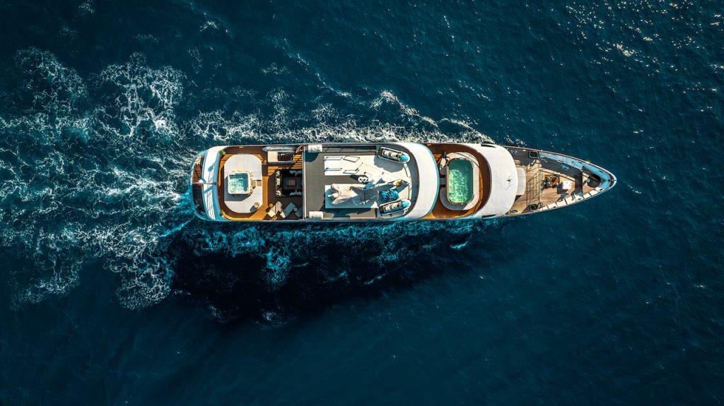 Aerial view of a luxurious yacht for charter sailing through deep blue ocean waters, leaving a trail of white waves behind. The yacht features sun loungers, pools, and elegant deck areas for relaxation.