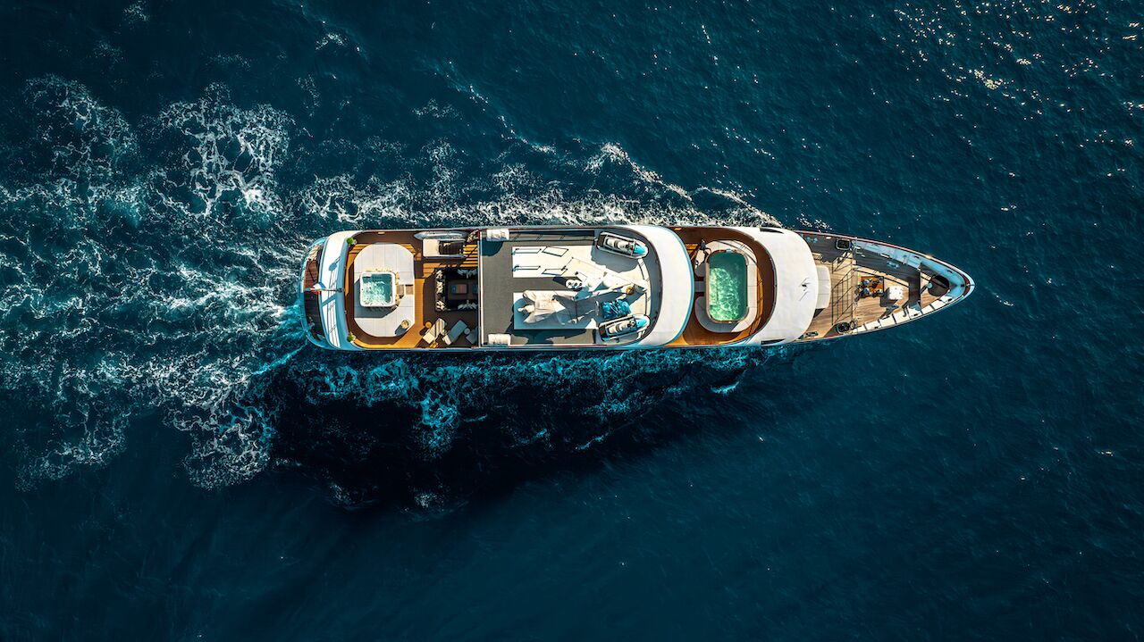 Aerial view of a luxurious yacht for charter sailing through deep blue ocean waters, leaving a trail of white waves behind. The yacht features sun loungers, pools, and elegant deck areas for relaxation.