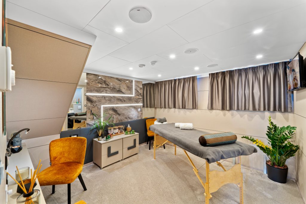A modern spa treatment room with a massage table, two orange chairs, and calming decor—perfect for unwinding after a day on a yacht charter. Enjoy neatly arranged towels, soothing products, and tranquil views through the large window with gray curtains.