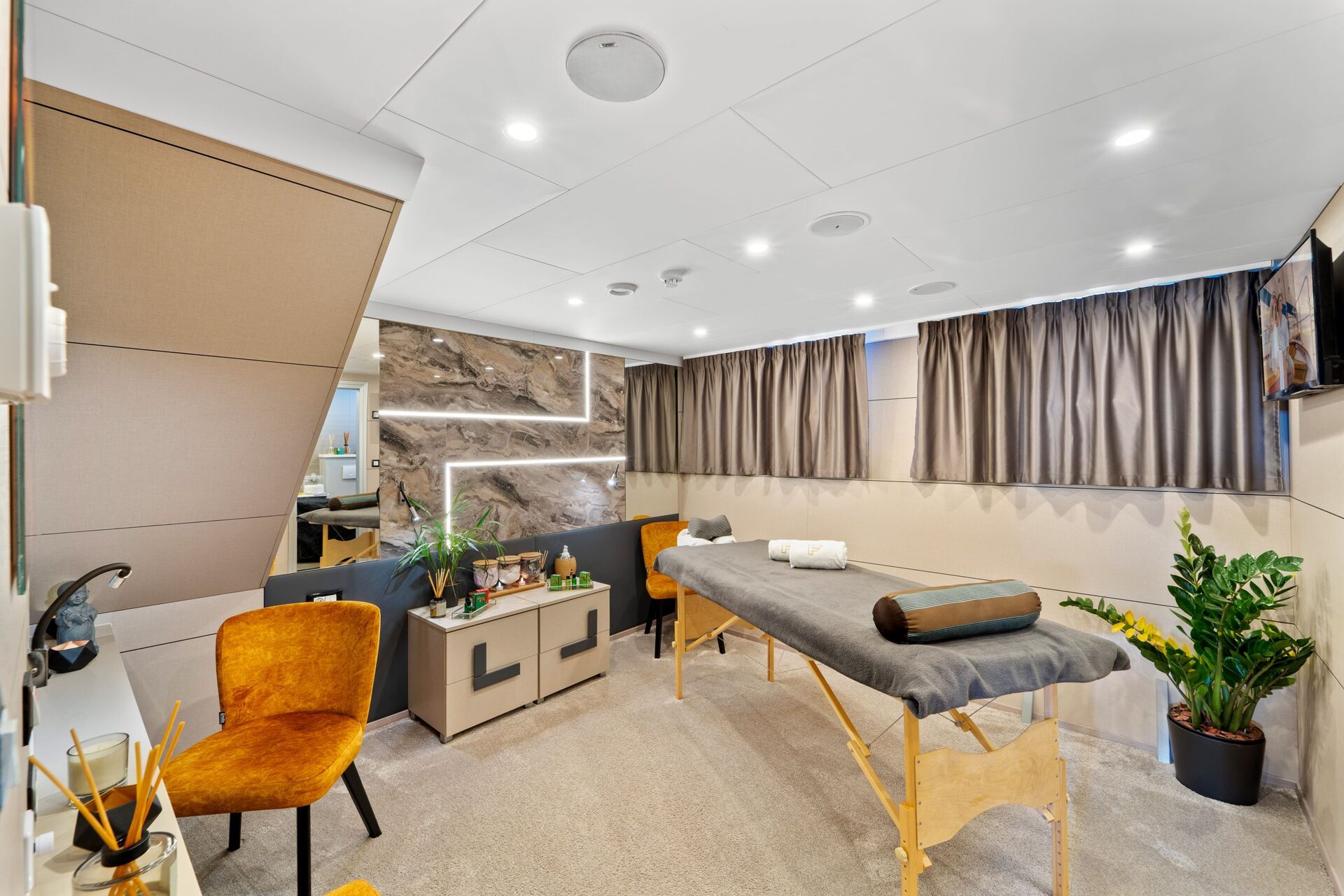 A modern spa treatment room with a massage table, two orange chairs, and calming decor—perfect for unwinding after a day on a yacht charter. Enjoy neatly arranged towels, soothing products, and tranquil views through the large window with gray curtains.