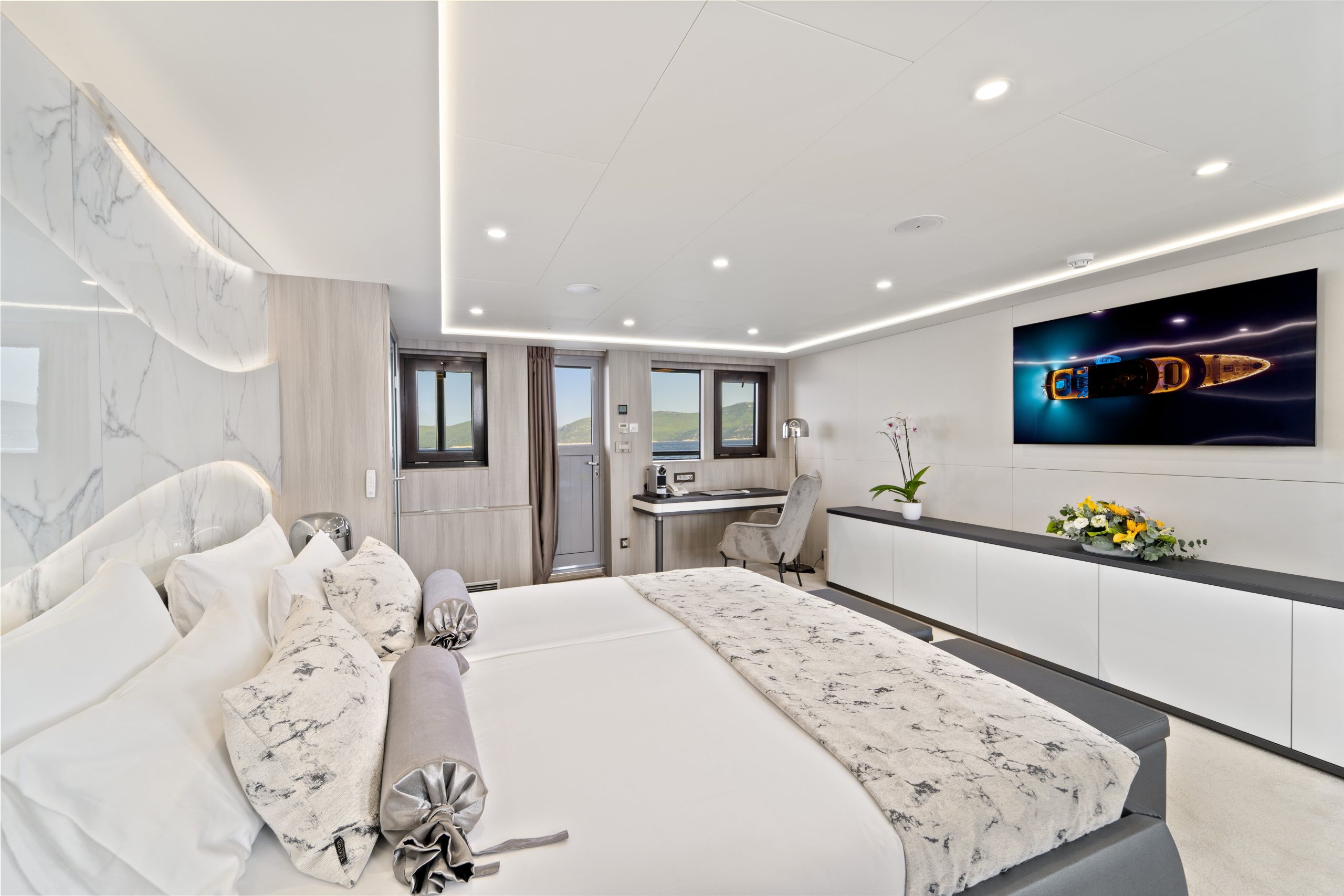 A modern bedroom features a large bed with white and marble-patterned bedding, a desk by two windows, a wall-mounted TV displaying a yacht charter, and minimalist decor with flowers and soft lighting.