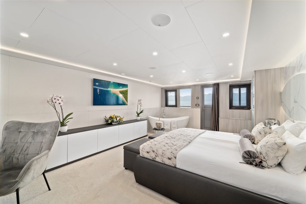 Modern, spacious bedroom with a large bed, elegant decor, two windows, a wall-mounted TV displaying yachts for charter gliding over the ocean, a gray chair, orchids, and a freestanding bathtub near the windows.
