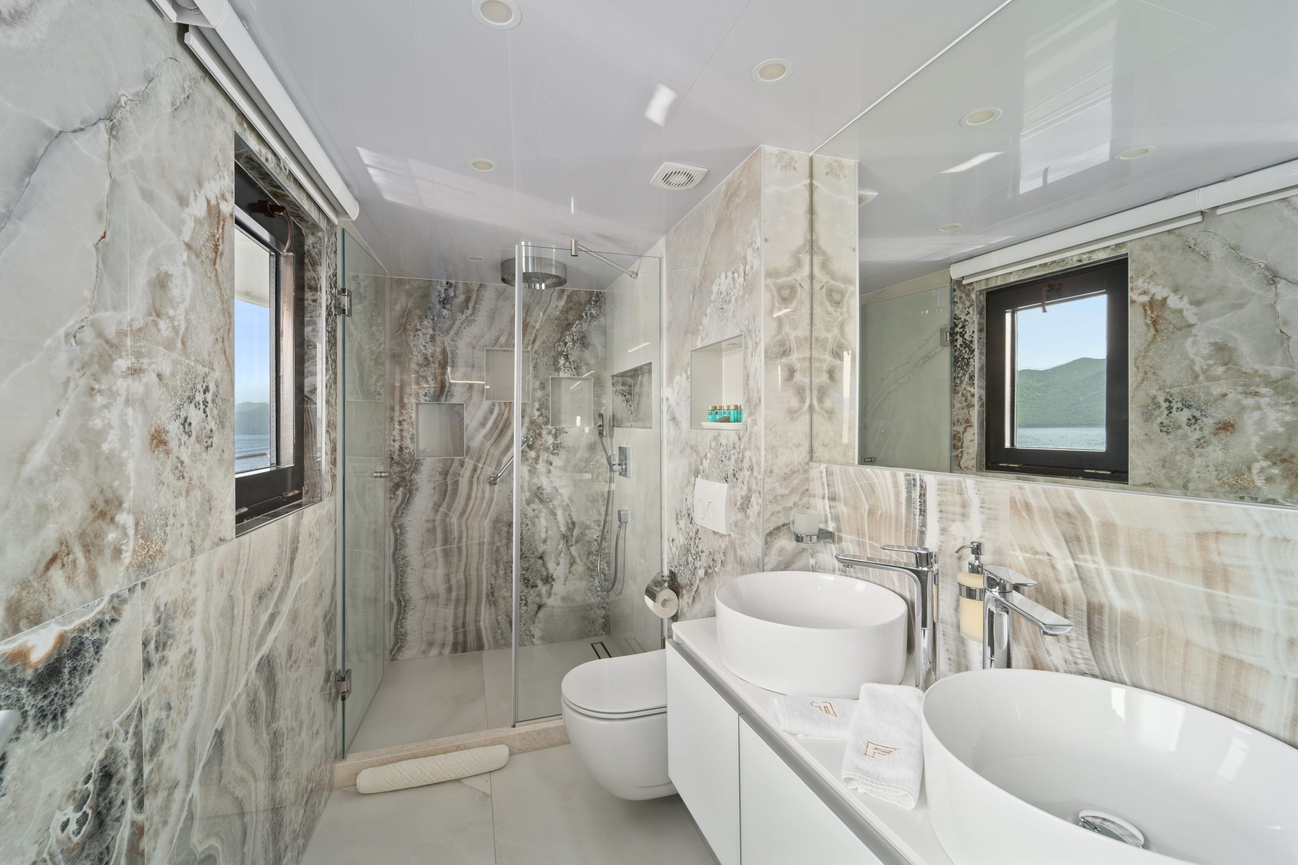 Modern bathroom with marble walls, two round vessel sinks on a white vanity, a glass-enclosed shower, large mirror, and neatly folded towels—perfect for luxury yachts for charter seeking comfort and style.