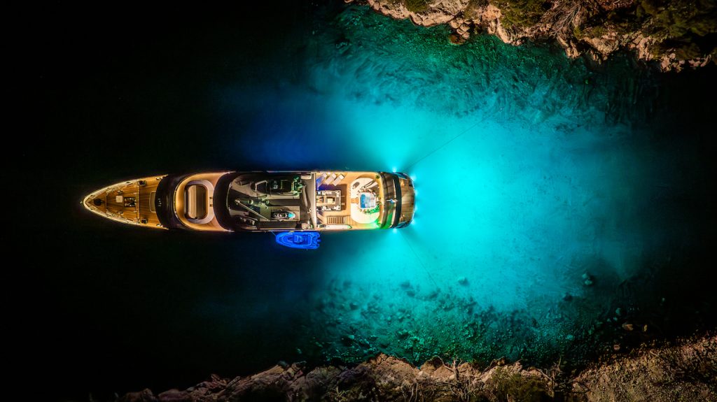 Aerial view of a yacht charter illuminated with bright blue underwater lights, anchored at night in clear water near a rocky shoreline.