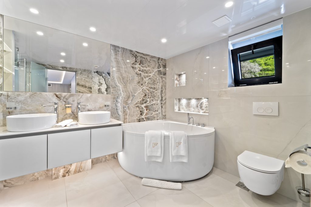 Modern bathroom with a large round bathtub, two vessel sinks on a long vanity, marble accents, and a window with a green landscape—reminiscent of the luxurious style found on premium yachts for charter. Towels, mirrors, and recessed lighting complete the look.