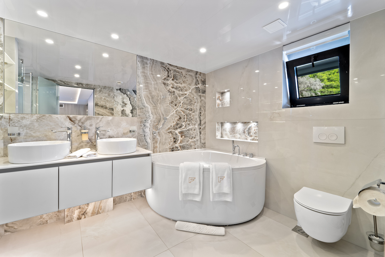 Modern bathroom with a large round bathtub, two vessel sinks on a long vanity, marble accents, and a window with a green landscape—reminiscent of the luxurious style found on premium yachts for charter. Towels, mirrors, and recessed lighting complete the look.