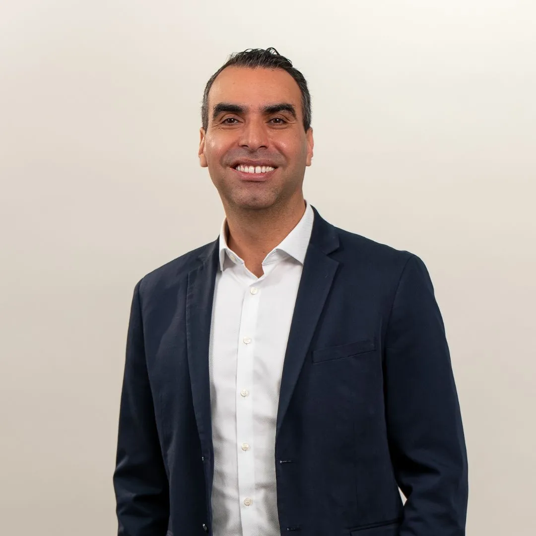 A man with dark hair wearing a navy blue suit jacket and white dress shirt smiles while standing against a plain light background.