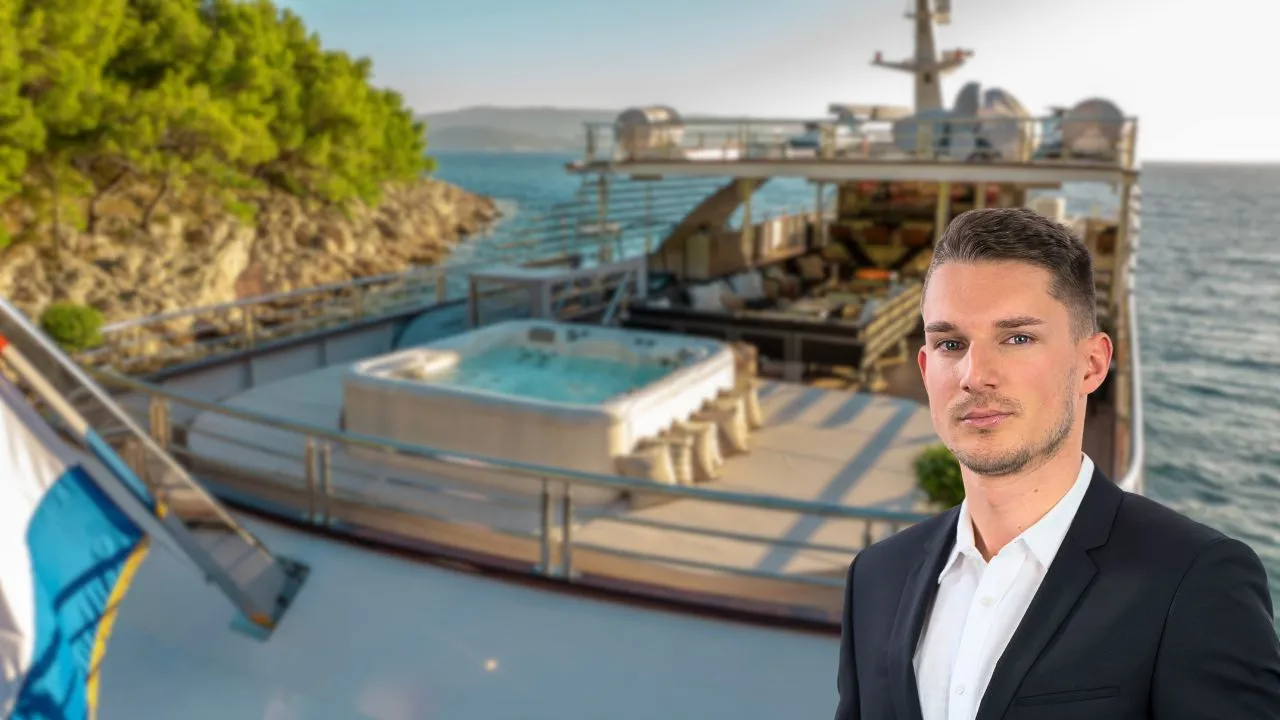 A man in a dark suit stands on the deck of a luxury yacht, enjoying the freedom of open water beside a hot tub, with a rocky, tree-covered shoreline and clear sunny skies in the background.