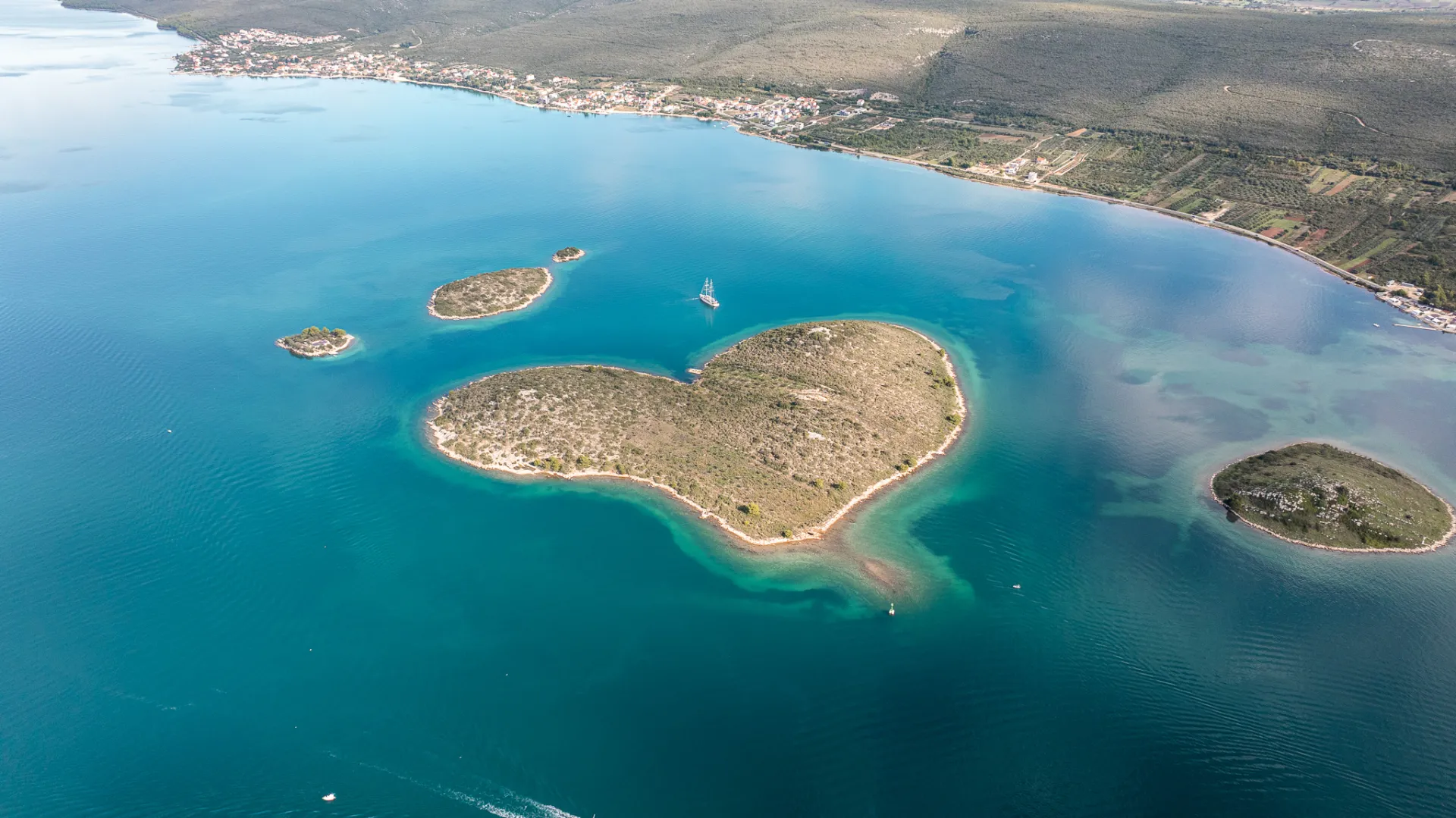 Aerial view of a heart-shaped island surrounded by clear blue water, with smaller islands nearby and a coastal town visible in the distance.