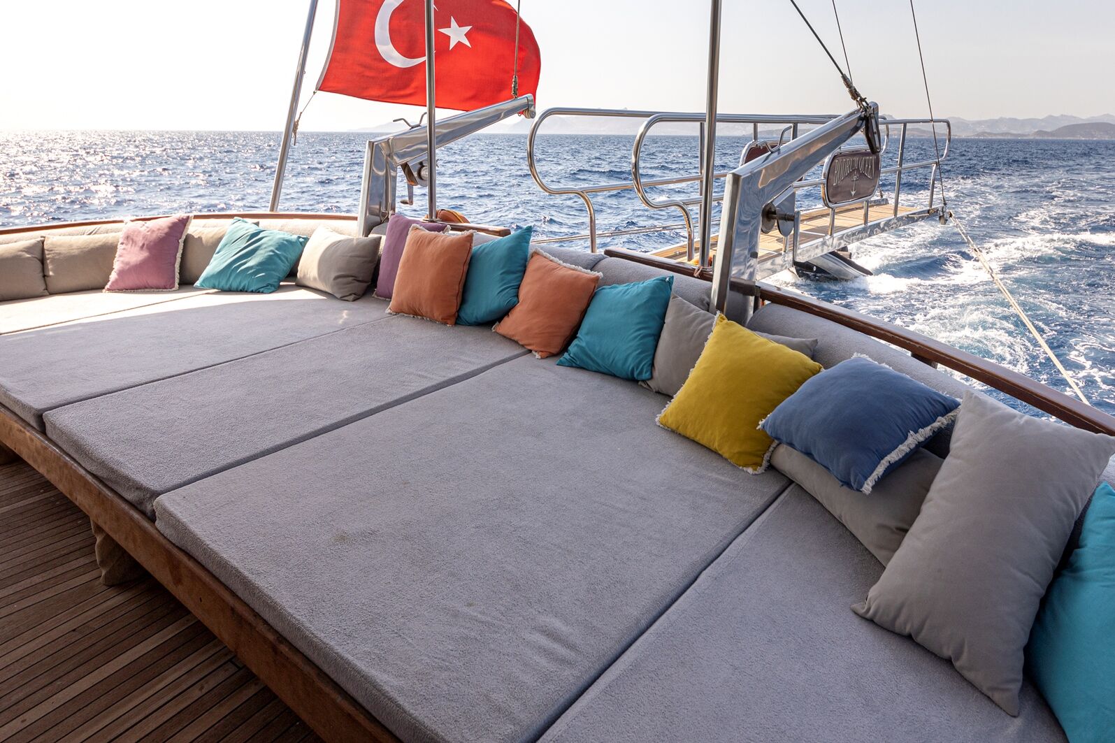 BODRUM QUEEN Large cushioned lounge area with colorful pillows on a yacht, a Turkish flag flying at the stern, and the boat sailing on a calm sea under a clear sky.