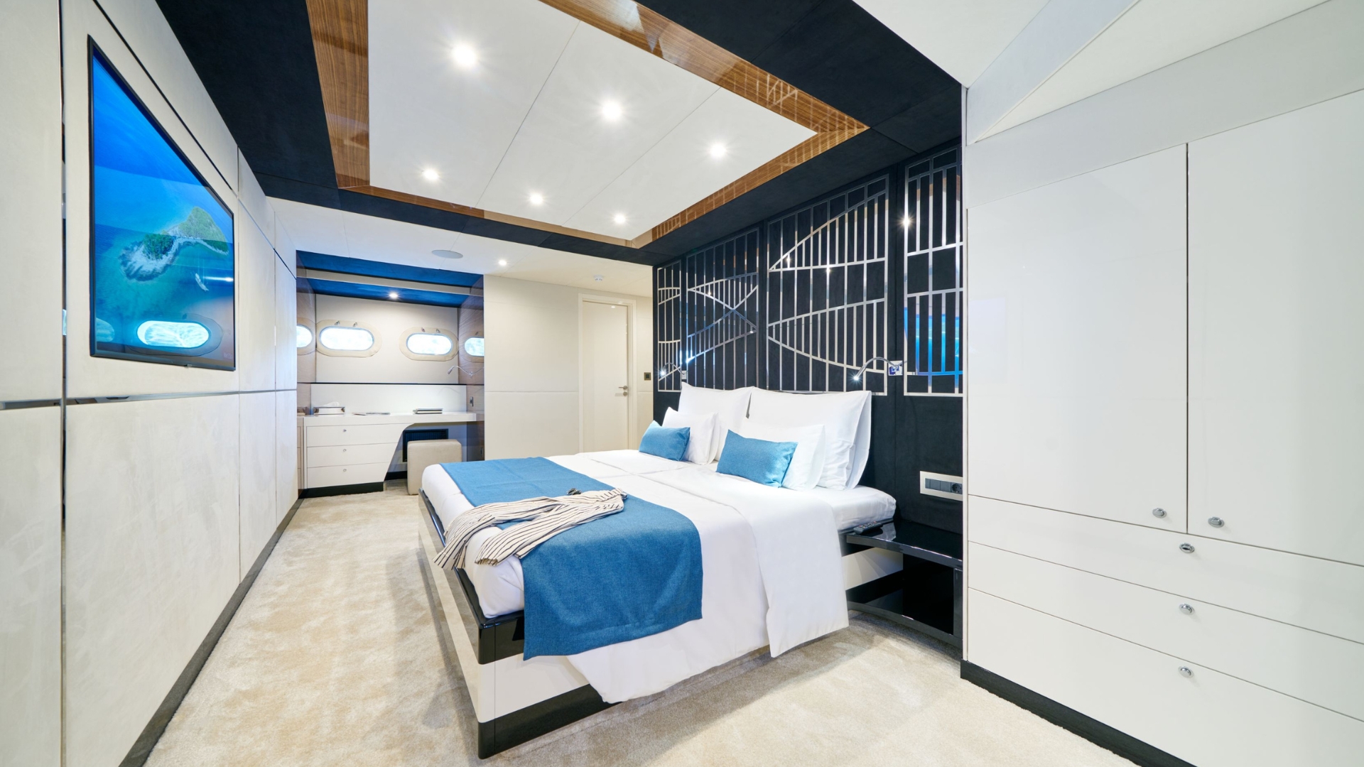 Modern bedroom with a large bed covered in white and blue linens, built-in wardrobe, light carpet, and contemporary decor. Windows and a TV displaying an ocean scene are visible. Room is brightly lit and sleekly designed.