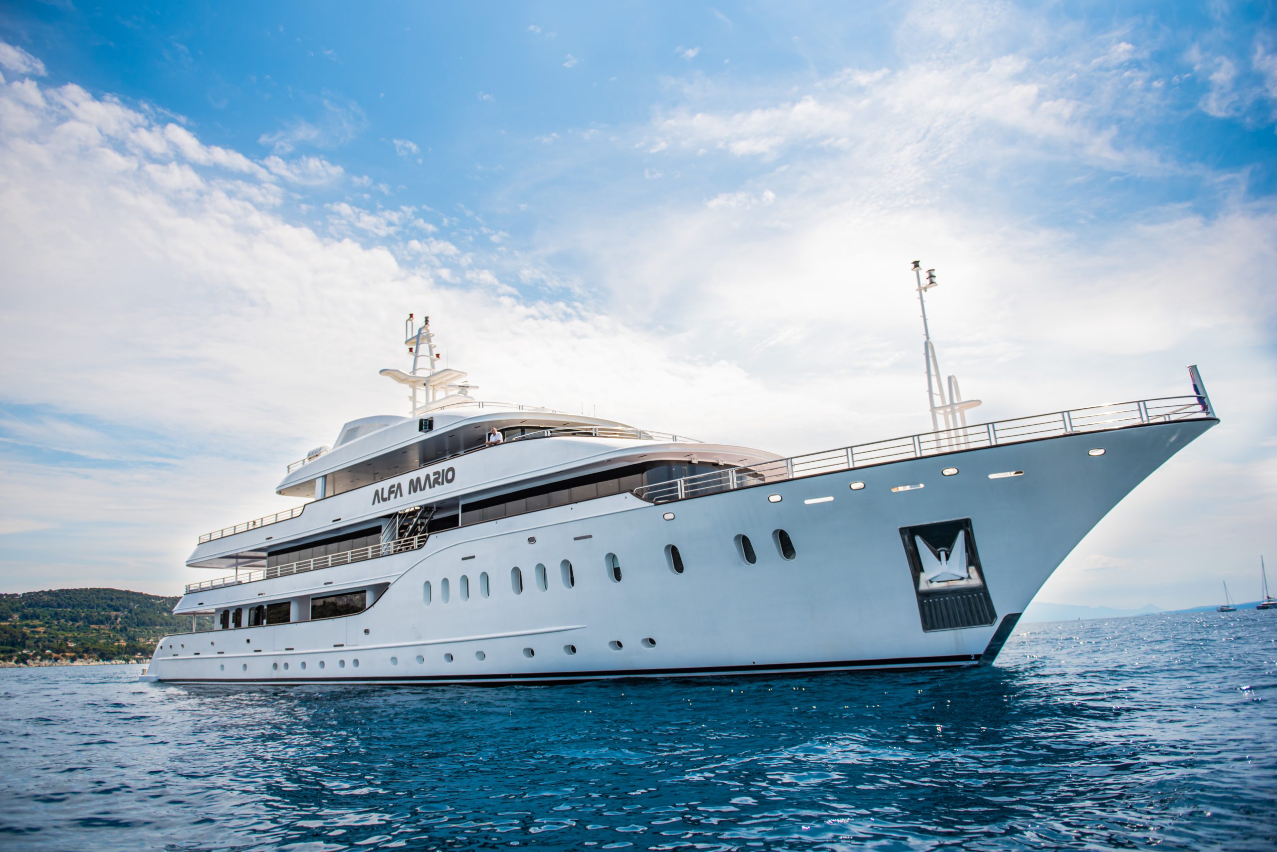 A large white luxury yacht named Alfa Mario is anchored on calm blue water under a partly cloudy sky, with coastal land visible in the background.