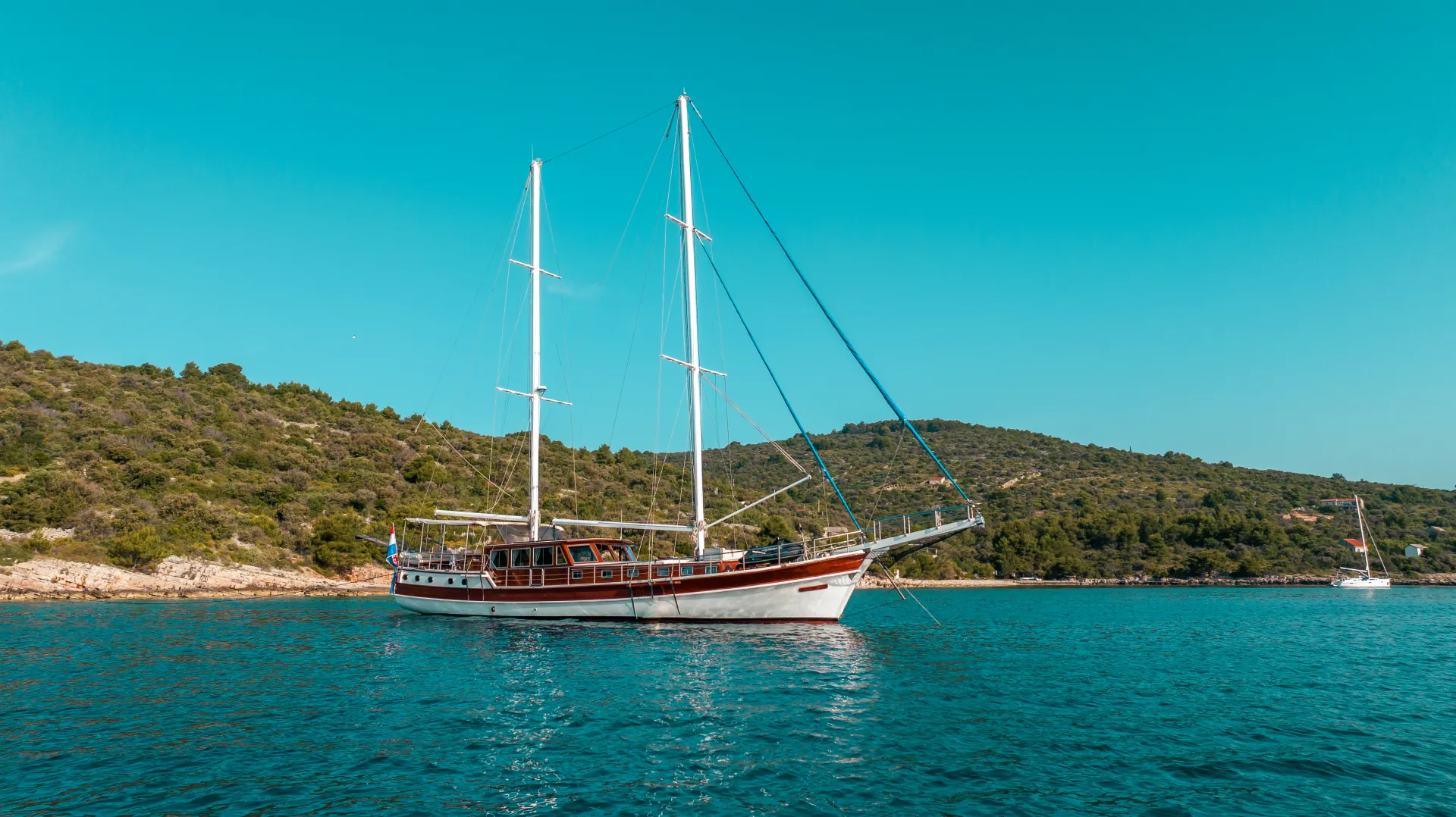 ANDI STAR A white sailboat with two masts floats on calm blue water near a green, hilly shoreline under a clear blue sky.