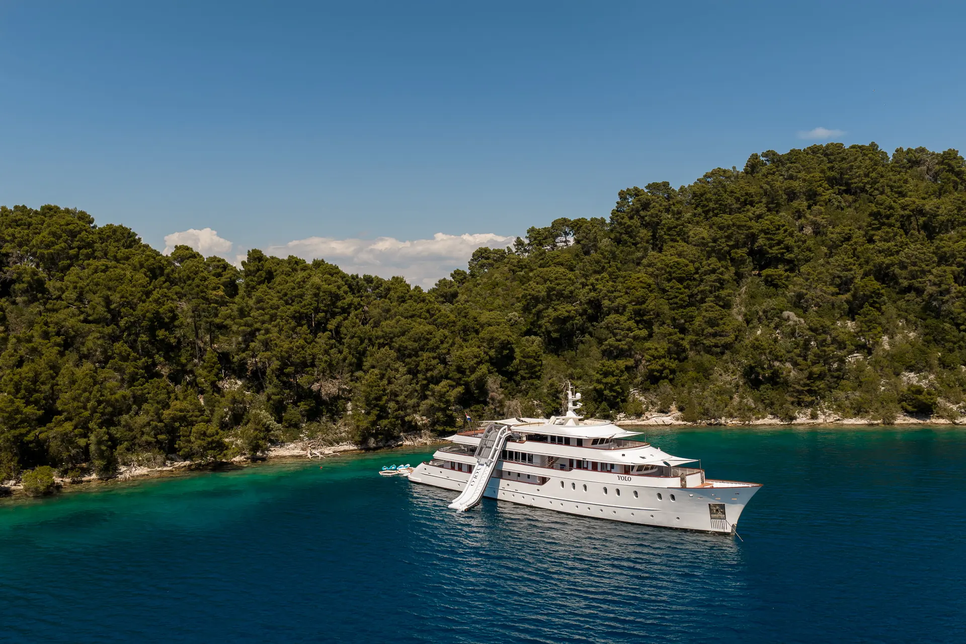 A large white yacht is anchored in clear blue-green water near a lush, forested shoreline under a sunny blue sky, with a slide leading from the yacht into the sea.