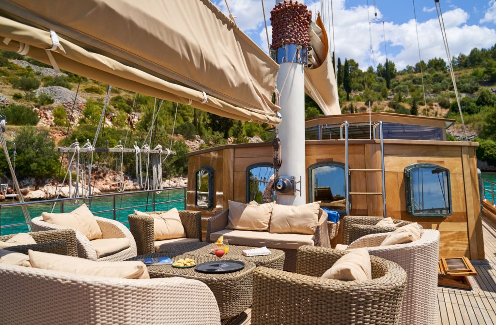 LIBRA A luxurious yacht deck with wicker chairs and beige cushions arranged around a small table, docked in turquoise water near a lush, green shoreline under a partly cloudy sky.