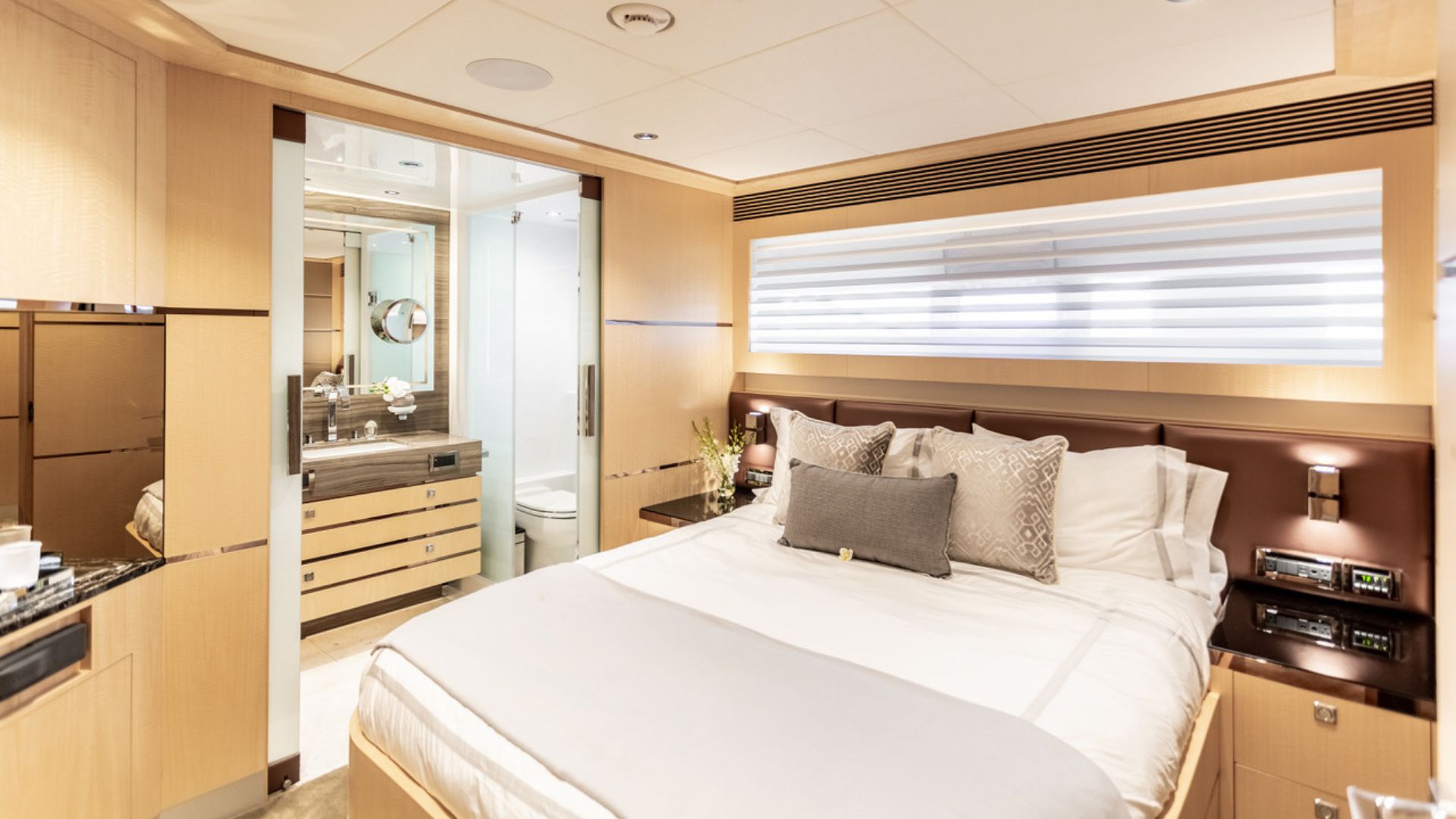 A modern yacht bedroom with light wood paneling, a neatly made bed with gray and white pillows, bedside tables, and an ensuite bathroom with a sink, mirror, and glass shower. Horizontal blinds cover the window.