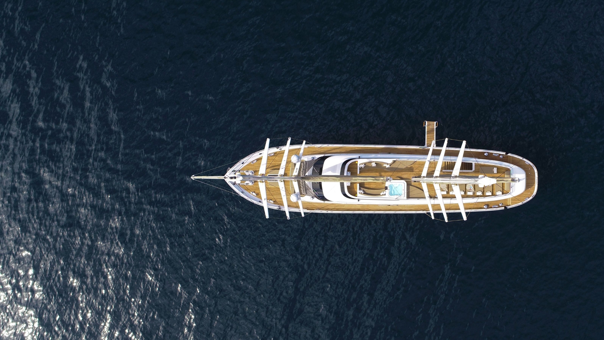 Aerial view of a large yacht with multiple decks floating on dark blue water, with sunlight reflecting off the surface beside the boat.