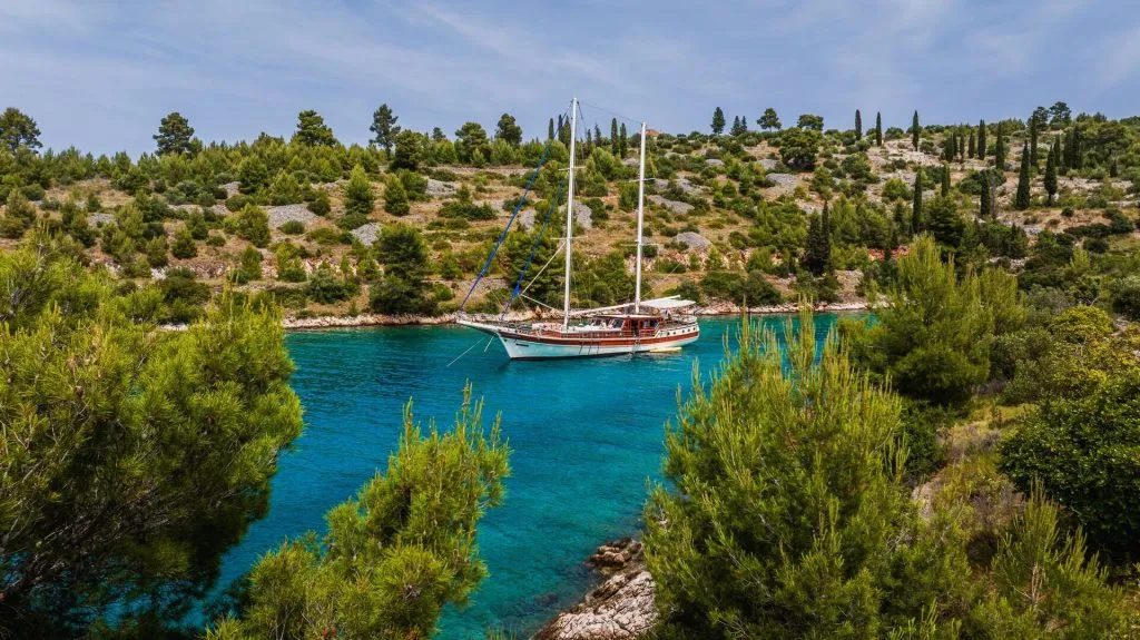 A sailboat with two masts floats on clear turquoise water, surrounded by lush green trees and hills under a partly cloudy sky.