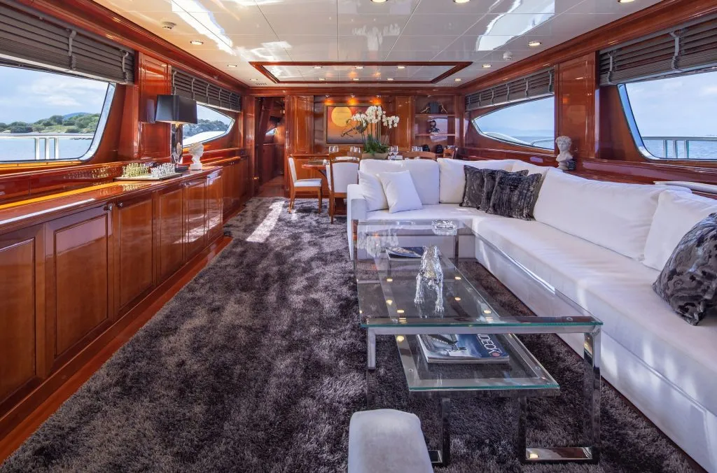 DIVINE Luxurious yacht interior with glossy wood paneling, large windows, a long white sectional sofa, glass coffee table, plush gray carpet, and elegant decor, overlooking the sea.