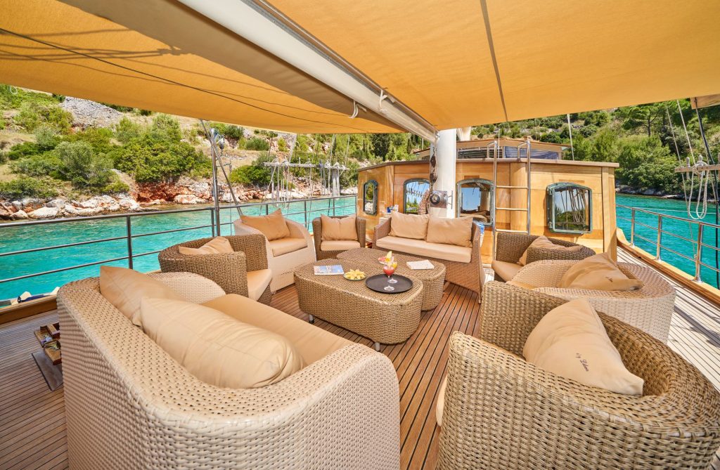 LIBRA Outdoor lounge area on a yacht with beige wicker sofas and chairs, a round table, and sun shade overhead. The yacht is anchored near clear turquoise water and lush green shoreline.