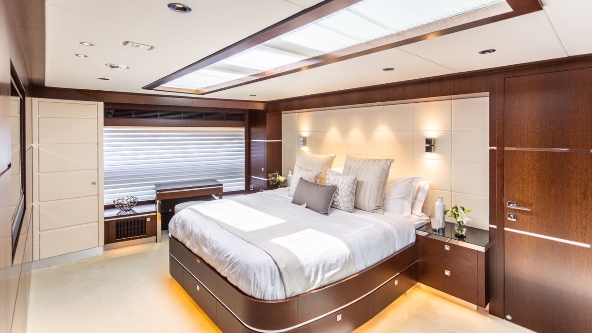 Luxurious modern bedroom with a large bed, white bedding, and accent pillows. The room features dark wood walls, built-in lighting, a window with blinds, and decorative plants, creating a bright and elegant atmosphere.