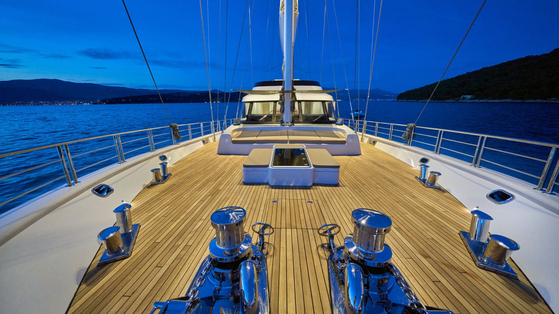 View from the deck of a luxury yacht at dusk, showing polished wood flooring, chrome fixtures, and calm blue water with distant hills under a twilight sky.