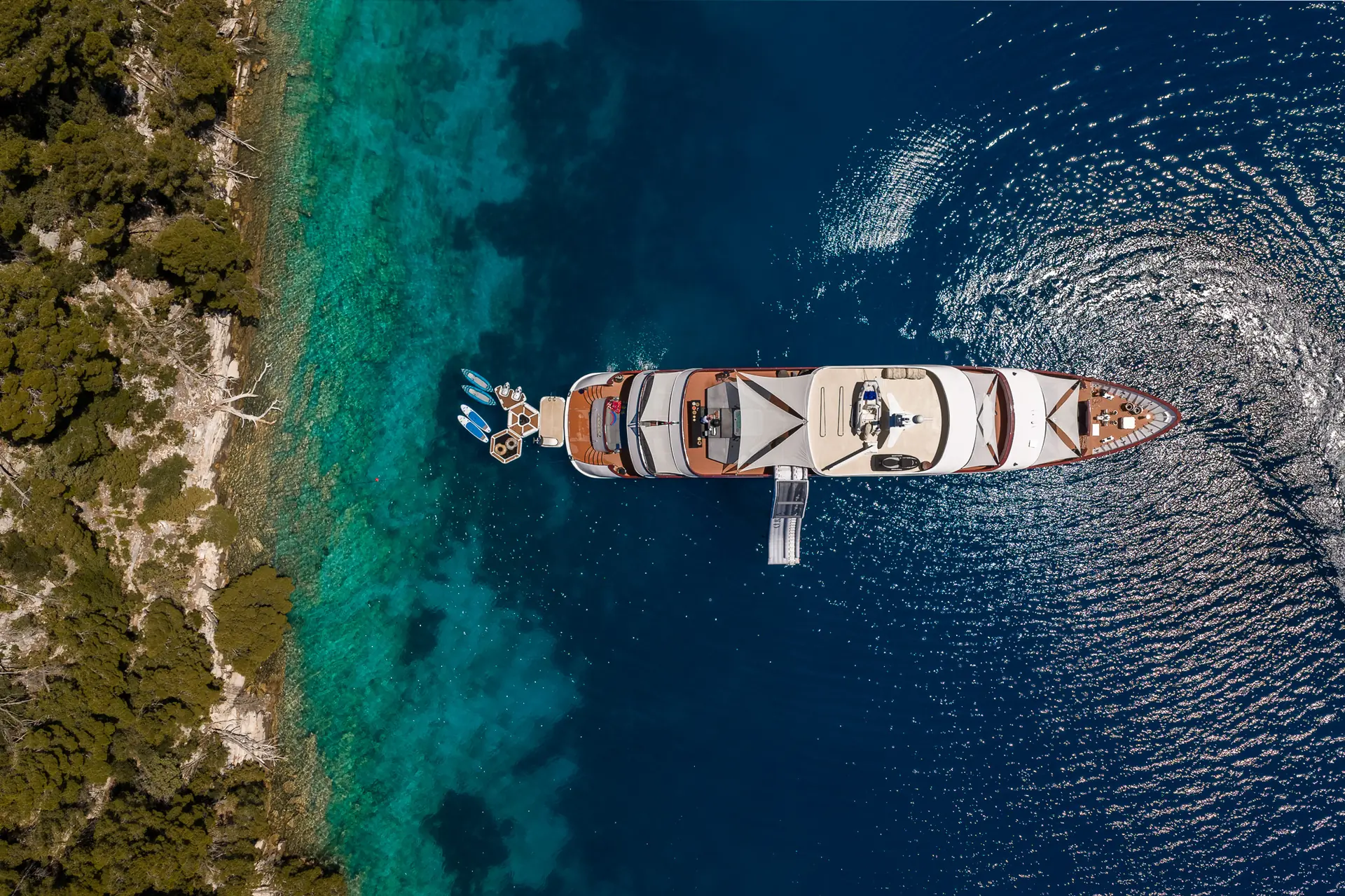 Aerial view of a large yacht anchored near a forested coastline with clear turquoise and deep blue water, people relaxing on the deck and swimming platforms extended over the water.