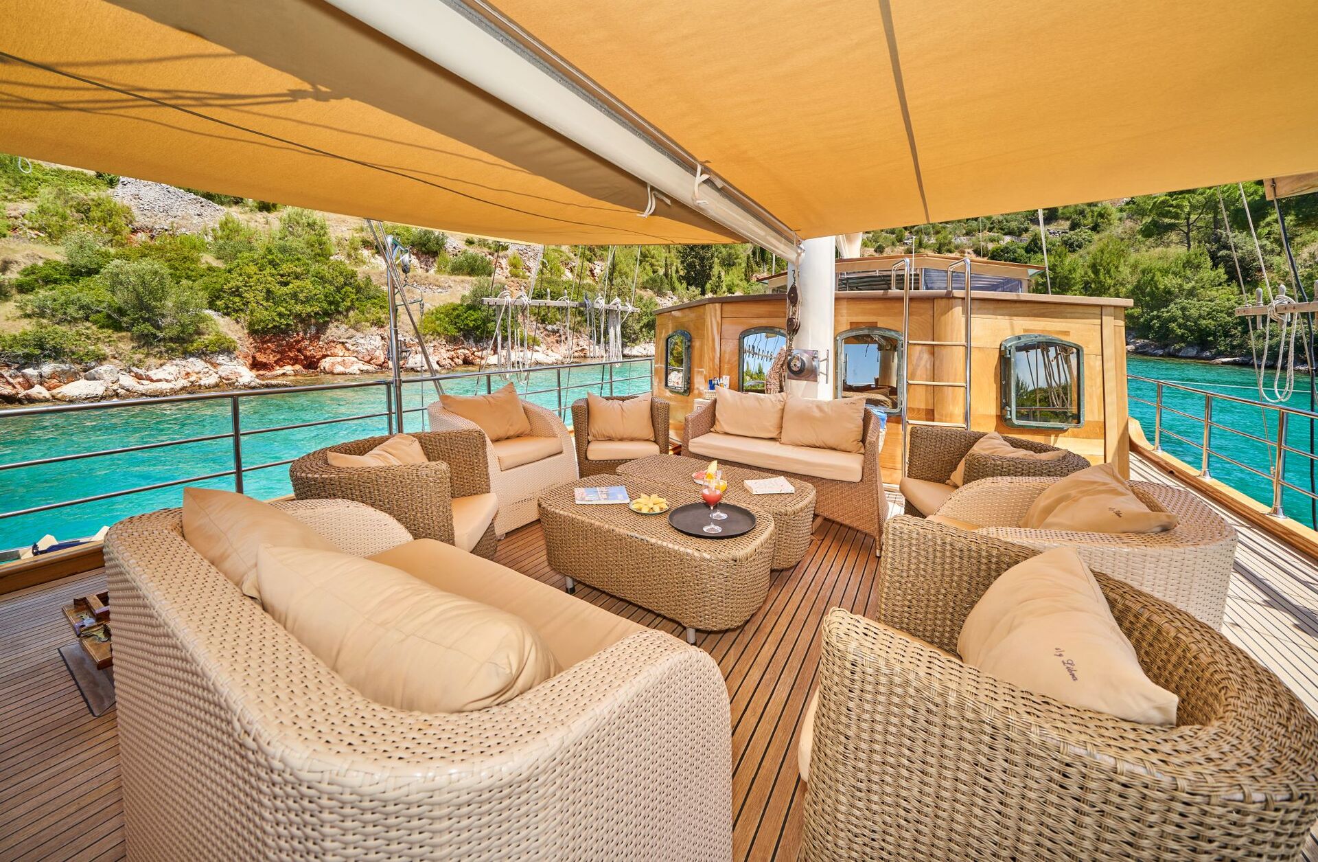 LIBRA Spacious yacht deck with wicker sofas and armchairs arranged around a table, overlooking turquoise water and a lush, rocky shoreline under a large tan canopy.