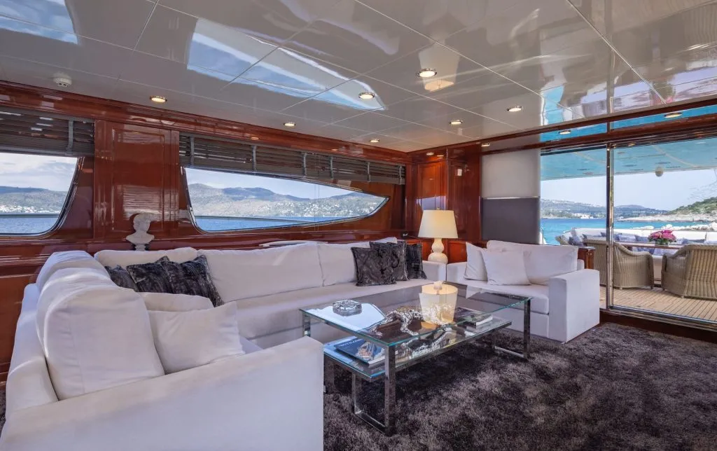 DIVINE uxurious yacht interior with white sofas, glass coffee table, and lamp on a dark rug. Large windows reveal scenic ocean and mountains, while an outdoor seating area is visible through a glass door.