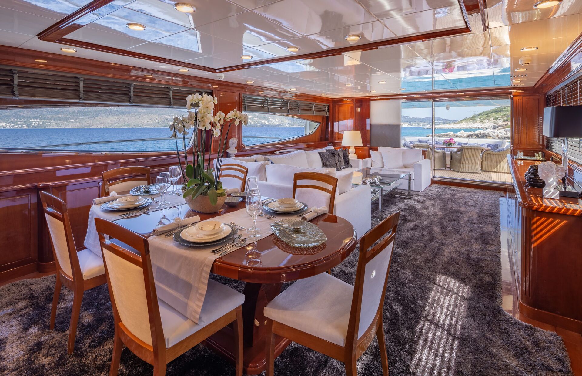 DIVINE Luxurious yacht interior with a set dining table, plush seating, large windows showcasing the ocean and coastline, elegant decor, and natural light filling the spacious, wood-accented room.