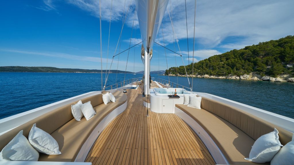 View from the deck of a luxury yacht with wooden flooring, cream-colored cushioned seating, and white pillows, sailing on calm blue water near a green, tree-lined coastline under a blue sky with scattered clouds.