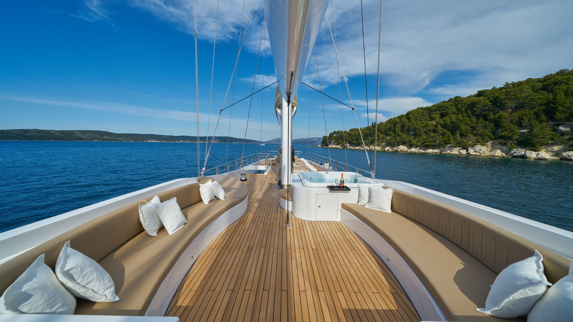 View from the deck of a luxury yacht with wooden flooring, cream-colored cushioned seating, and white pillows, sailing on calm blue water near a green, tree-lined coastline under a blue sky with scattered clouds.