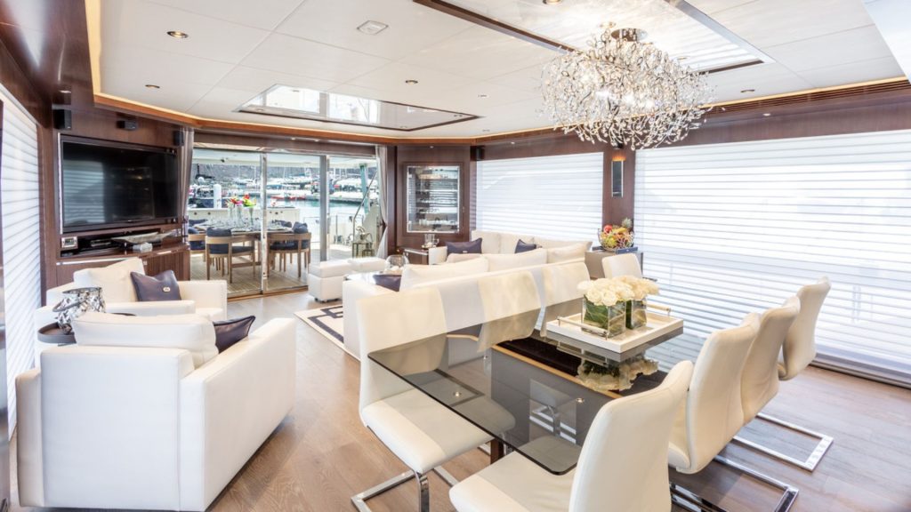 A luxurious yacht interior with white leather sofas, a glass dining table, elegant lighting, large windows with blinds, and a view of a marina through sliding glass doors at the back.