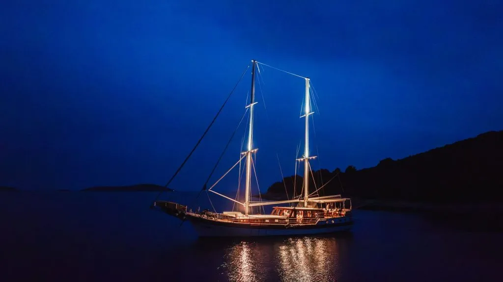 A sailboat with illuminated masts and deck lights floats on calm water at night, surrounded by darkness and silhouetted landforms in the background.