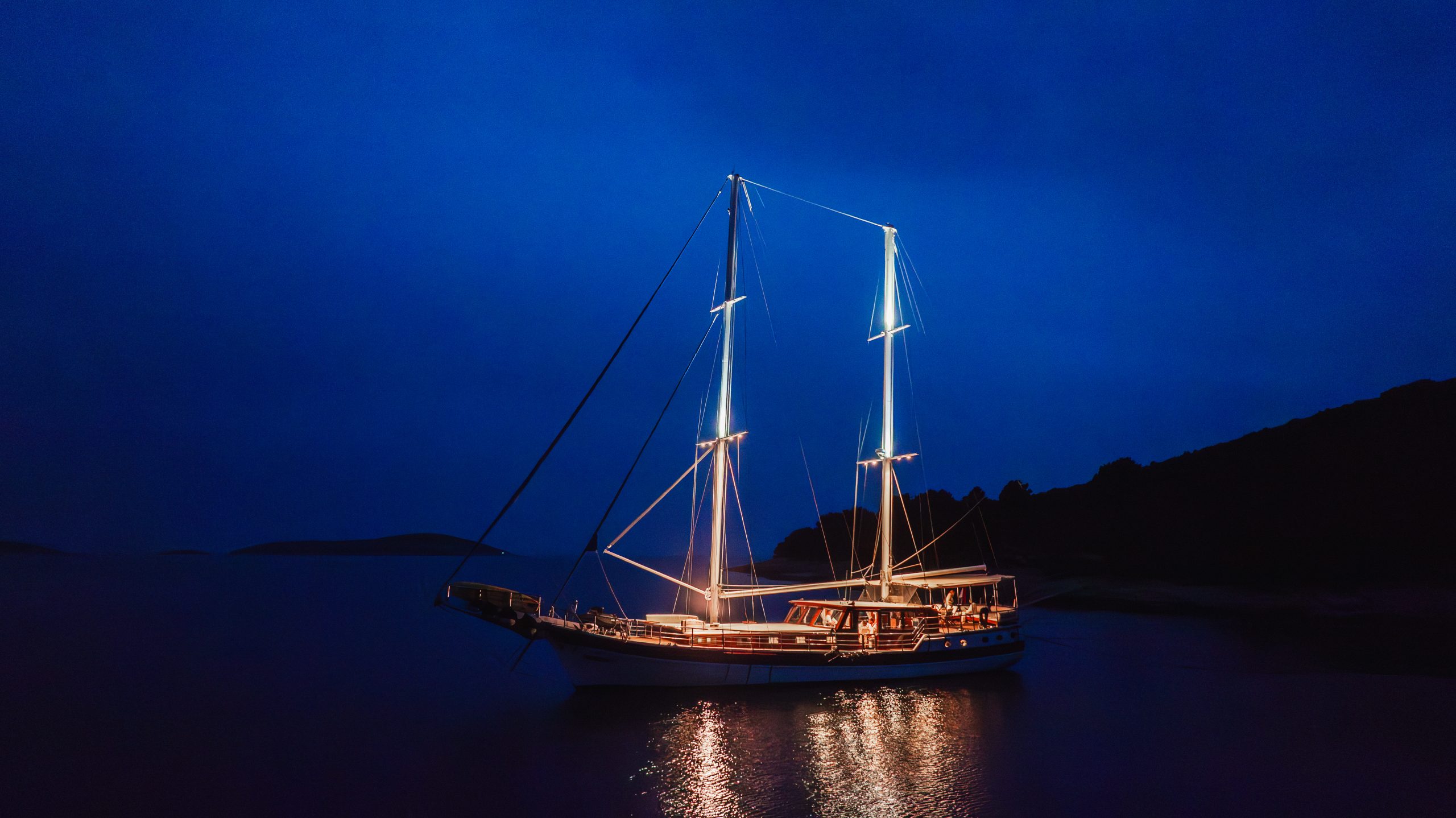 A sailboat with illuminated masts and deck lights floats on calm water at night, surrounded by darkness and silhouetted landforms in the background.