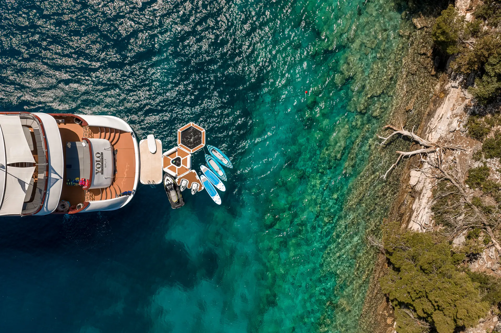 Aerial view of a yacht anchored near a rocky shoreline, with floating platforms and paddleboards on clear turquoise water, surrounded by greenery and a few fallen branches.