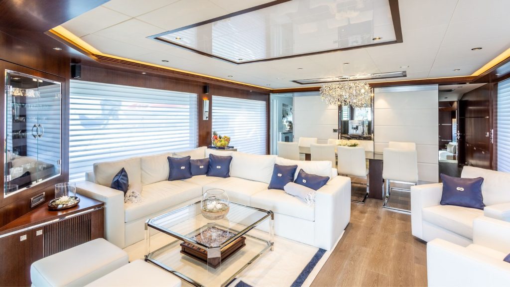 Modern yacht lounge with white leather sofas, blue accent pillows, a glass coffee table, wood panel walls, large windows with blinds, and a dining area with a crystal chandelier in the background.