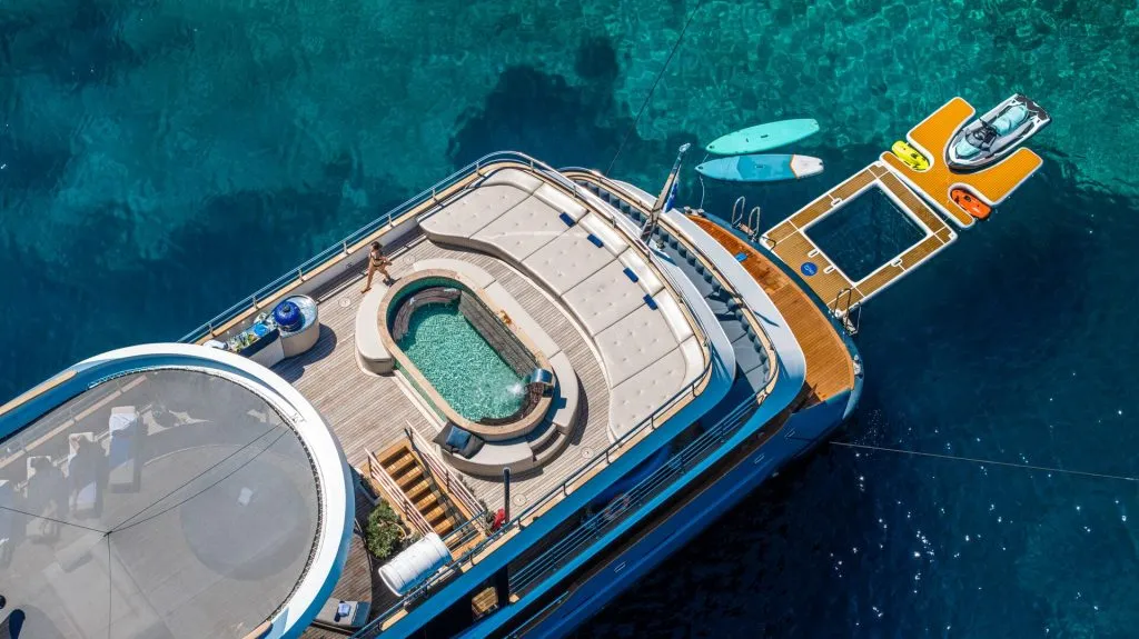 Aerial view of a luxury yacht with a pool on deck, anchored in clear blue water. Paddleboards, a jet ski, and an inflatable platform float nearby.