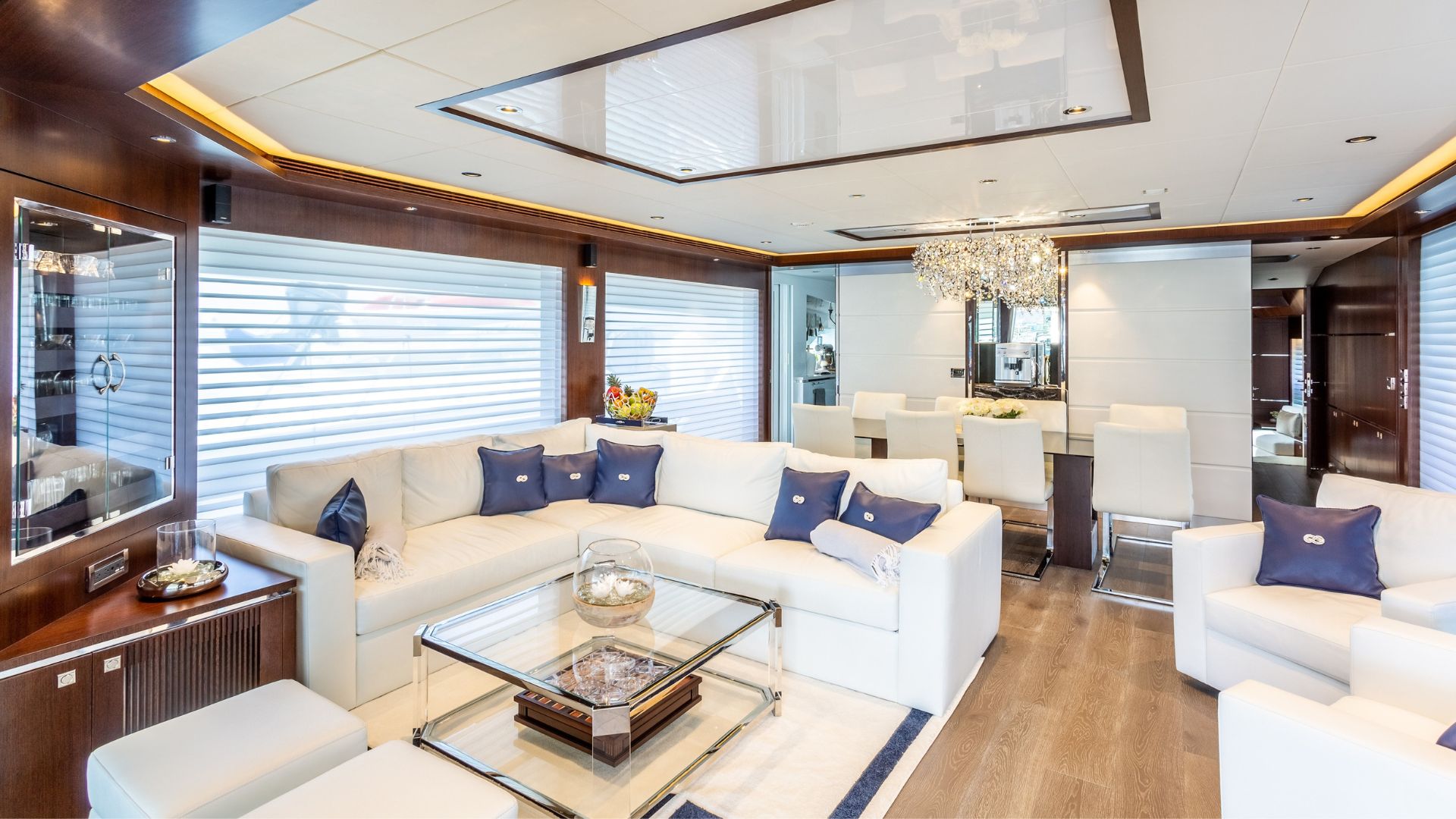 Modern yacht lounge with white leather sofas, blue accent pillows, a glass coffee table, wood panel walls, large windows with blinds, and a dining area with a crystal chandelier in the background.