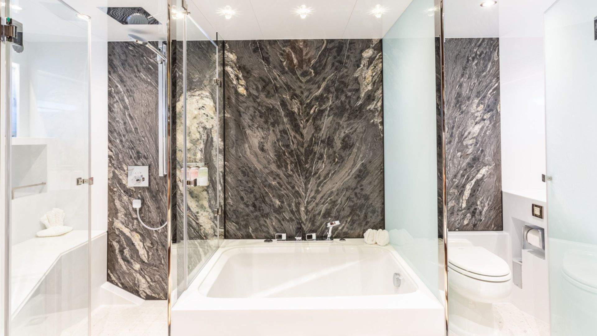 A modern bathroom featuring a large soaking tub surrounded by glass panels, marble-patterned walls, a walk-in shower on the left, and a toilet on the right. The space is bright and sleek with white and gray tones.