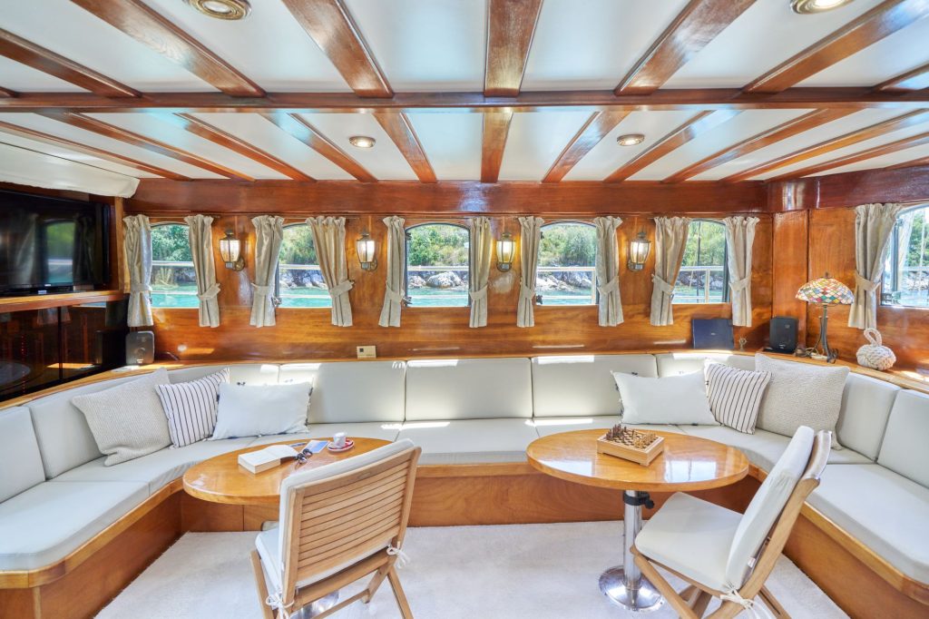 LIBRA A luxurious yacht interior with wooden paneling, cream-colored cushioned seating, striped throw pillows, round tables, a chess set, and large windows offering views of water and greenery.