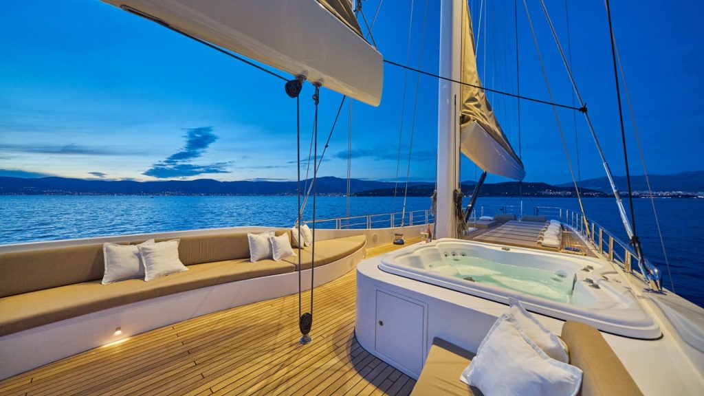 A luxury yacht deck at sunset features a built-in jacuzzi, cushioned seating with pillows, and a scenic view of calm water and distant mountains under a clear blue sky.